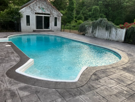 A kidney-shaped pool with blue water and gray stamped concrete patio, small white building in background.
