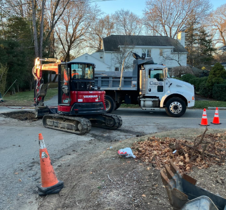 An excavator and dump truck on a street next to a hole. Two orange traffic cones are present.