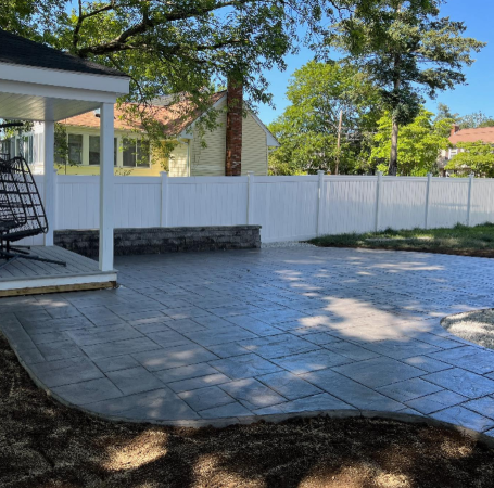 Backyard patio with patterned gray pavers, white fence, and small wall.