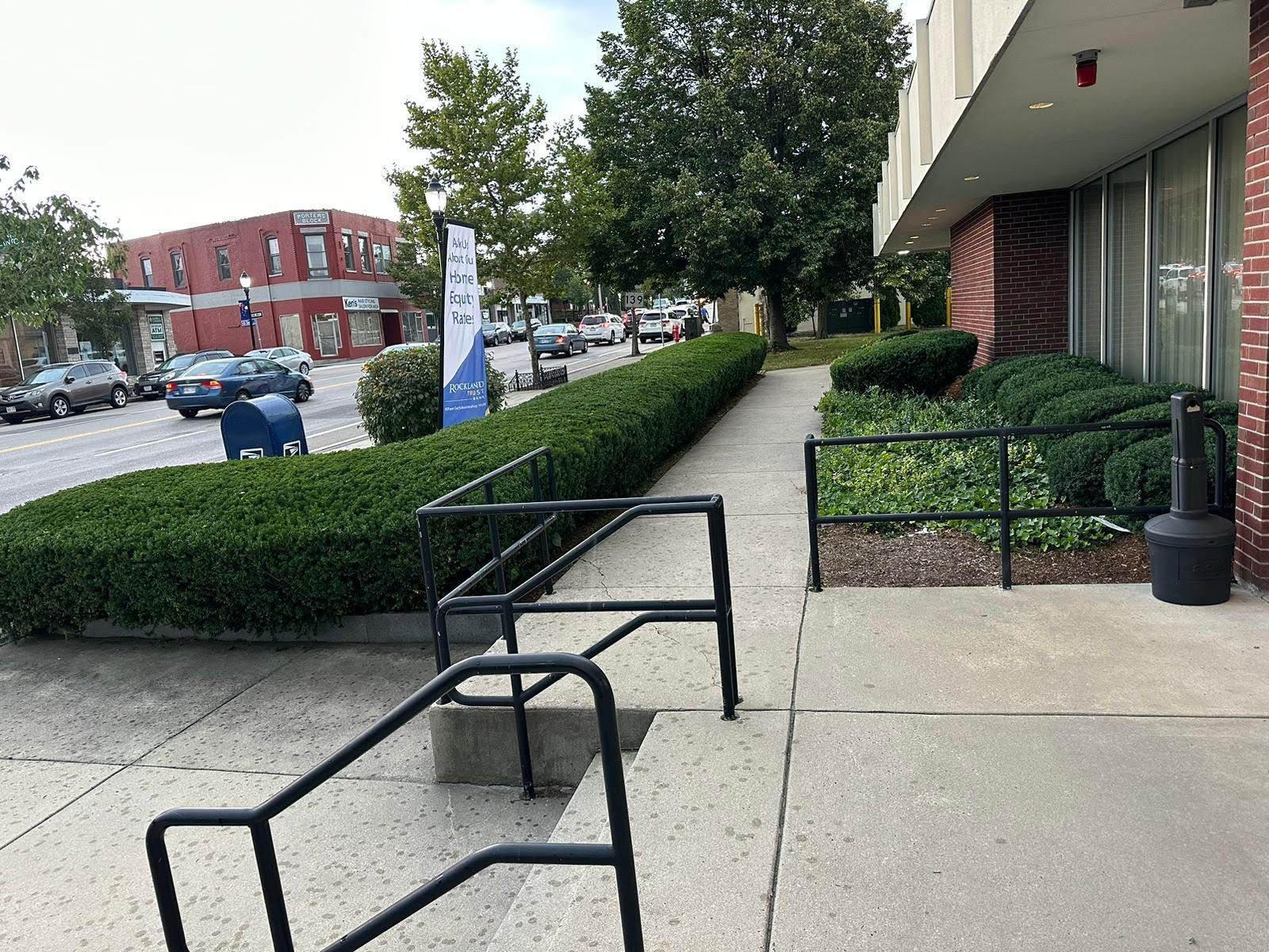 Exterior view of a building with a ramp and steps. A line of bushes separates the sidewalk from the street. Cars are visible.