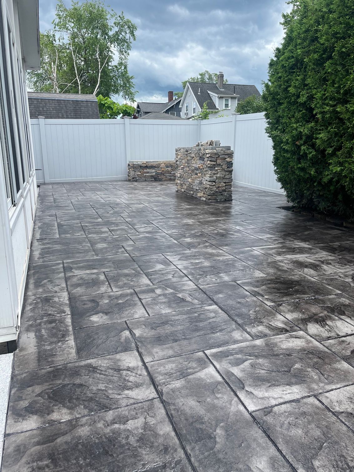 Gray stone patio with stone structure, white fence, and green foliage.