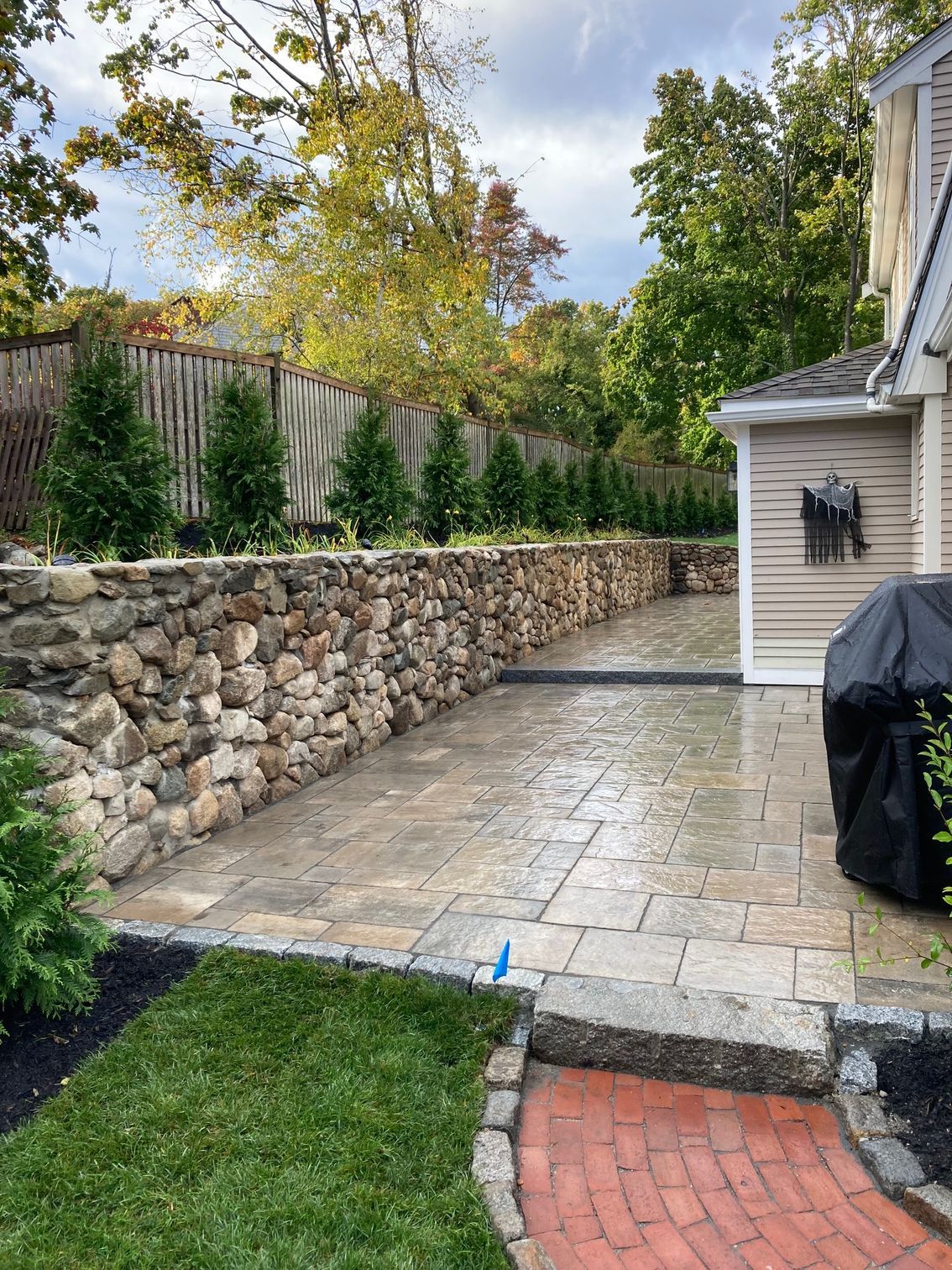 Stone patio with a stone wall and small evergreens along a wooden fence; a house is to the right.