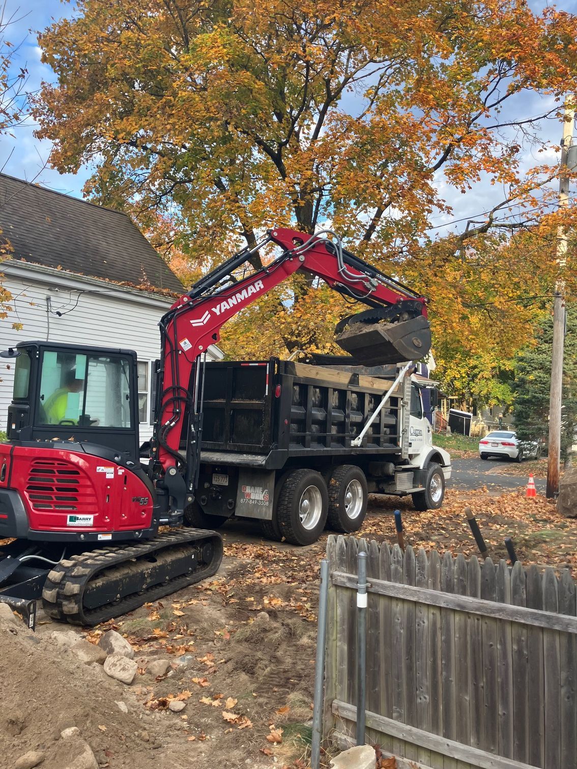 Red excavator loading a dump truck with dirt, trees with fall foliage in background.