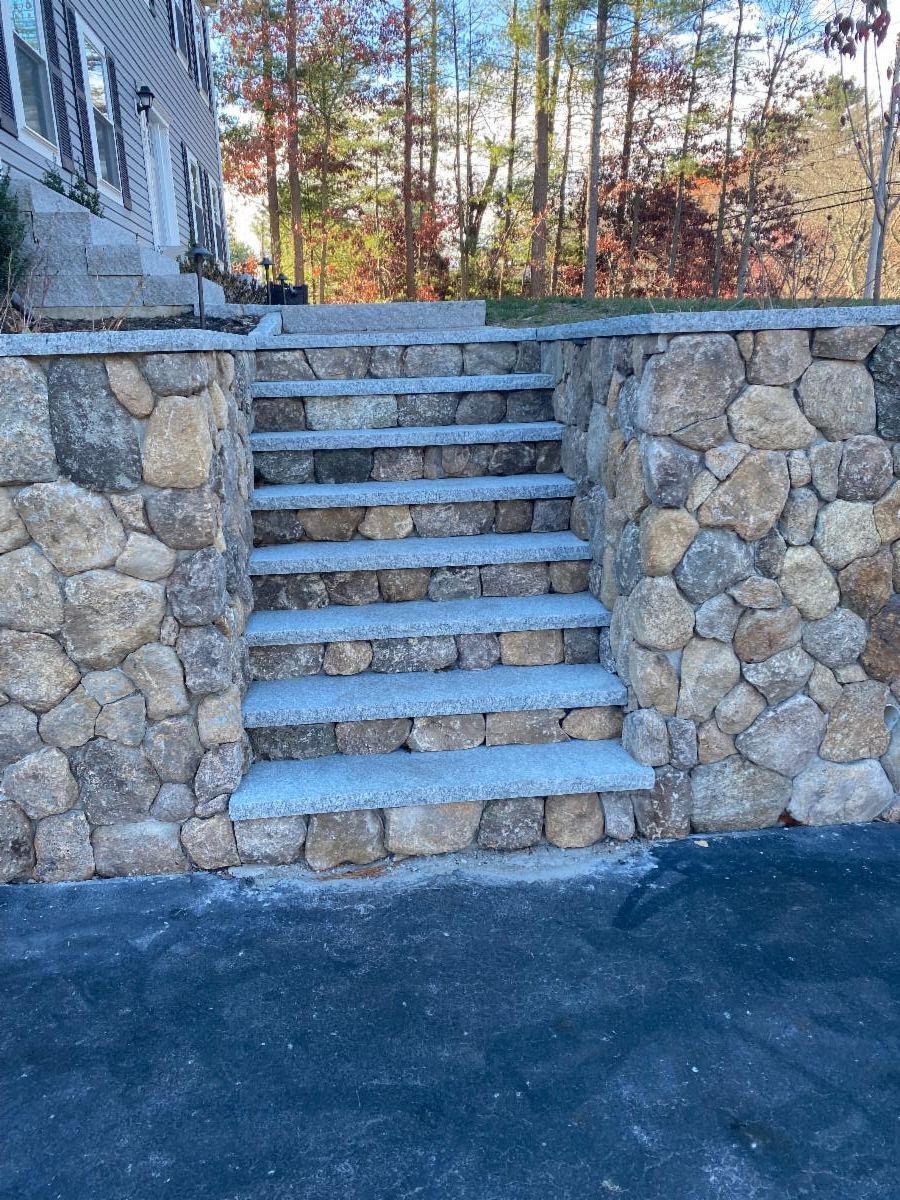 Stone steps built into a retaining wall. The steps are light gray, and the wall is made of brown stones.