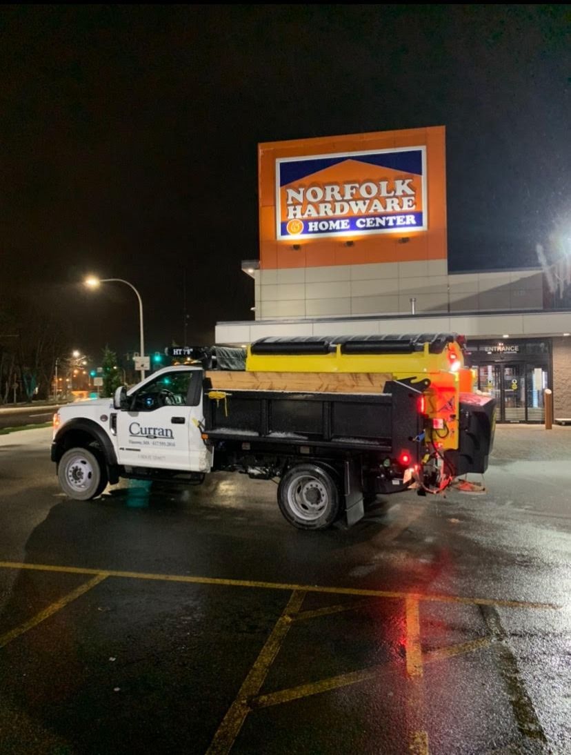 White work truck with a yellow salt spreader in front of a Norfolk Hardware store at night.