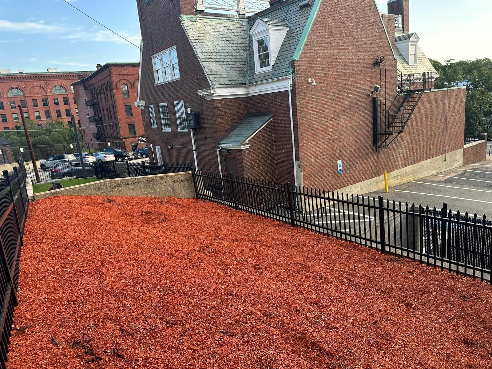 Red mulch-covered area with a black fence, adjacent to a brick building and a street with parked vehicles.