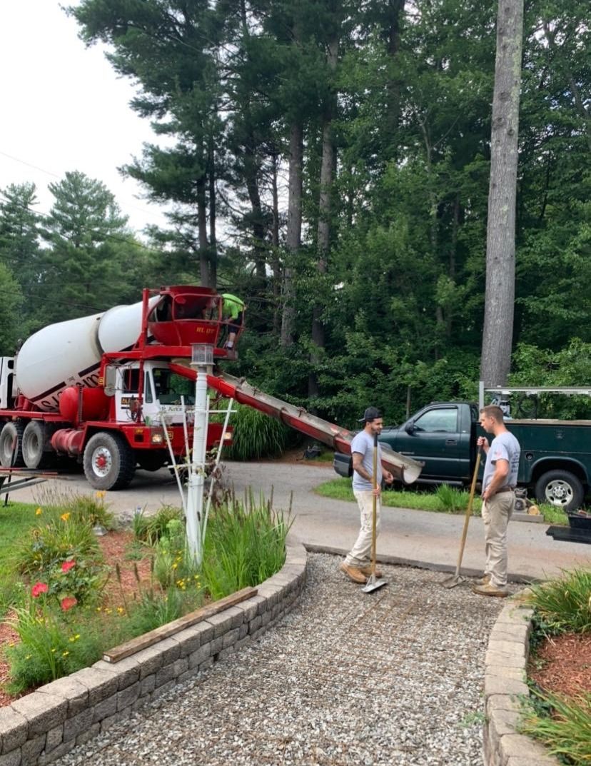 Concrete truck pouring cement onto gravel pathway; workers level it with tools. Outdoors, near trees.