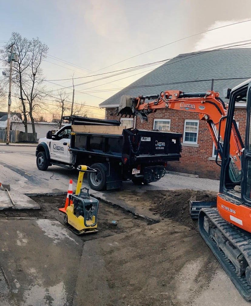 Construction site: excavator loading dump truck on a street. Compactor and orange cone in foreground. Buildings and sky in background.