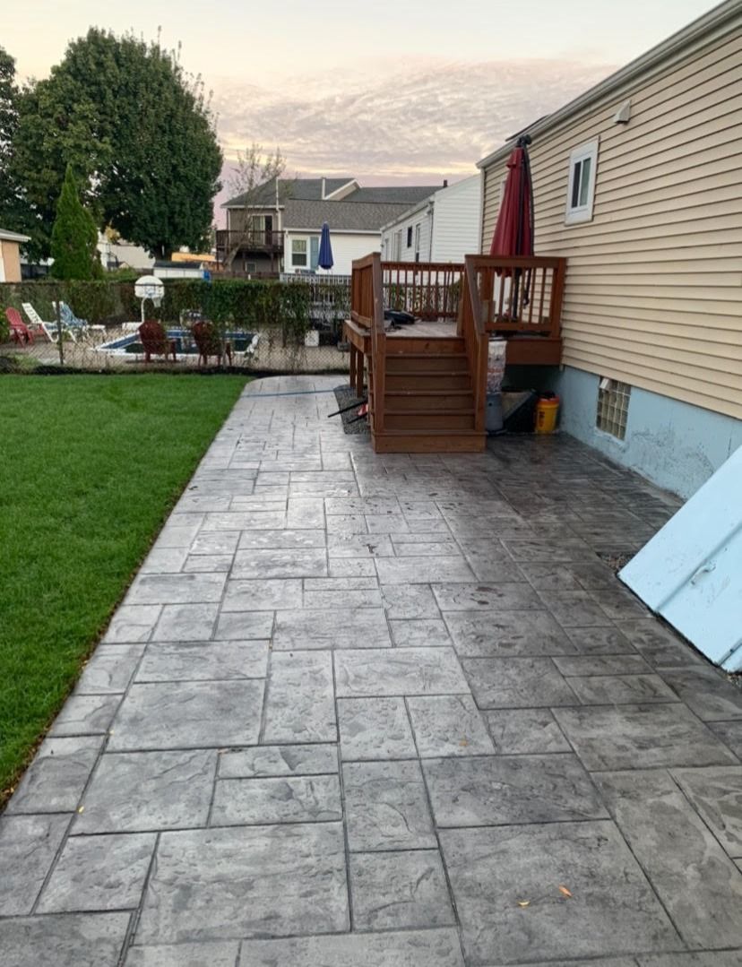 Backyard patio with stamped concrete, adjacent to a wooden deck. Green lawn, beige house, and overcast sky.