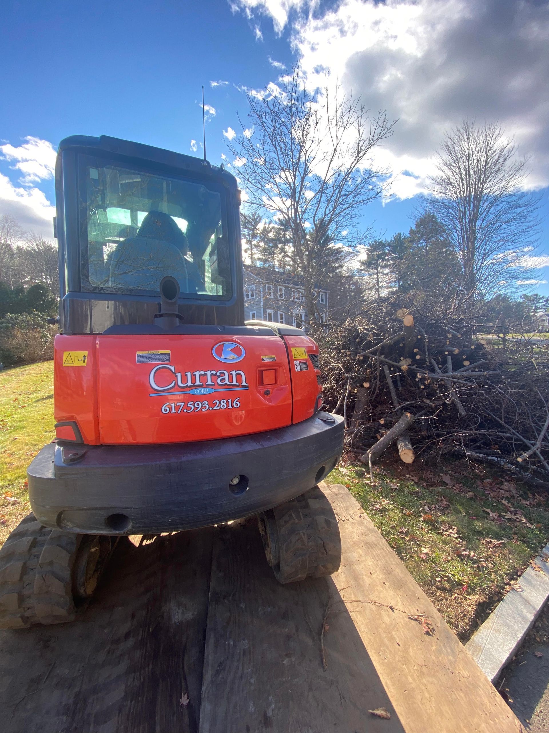 Orange Kubota compact excavator on wooden beams near a pile of branches under a cloudy sky.