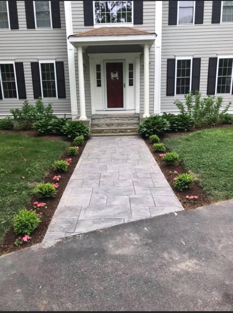 Stone walkway leading to a house with a red door, flanked by greenery and bushes.