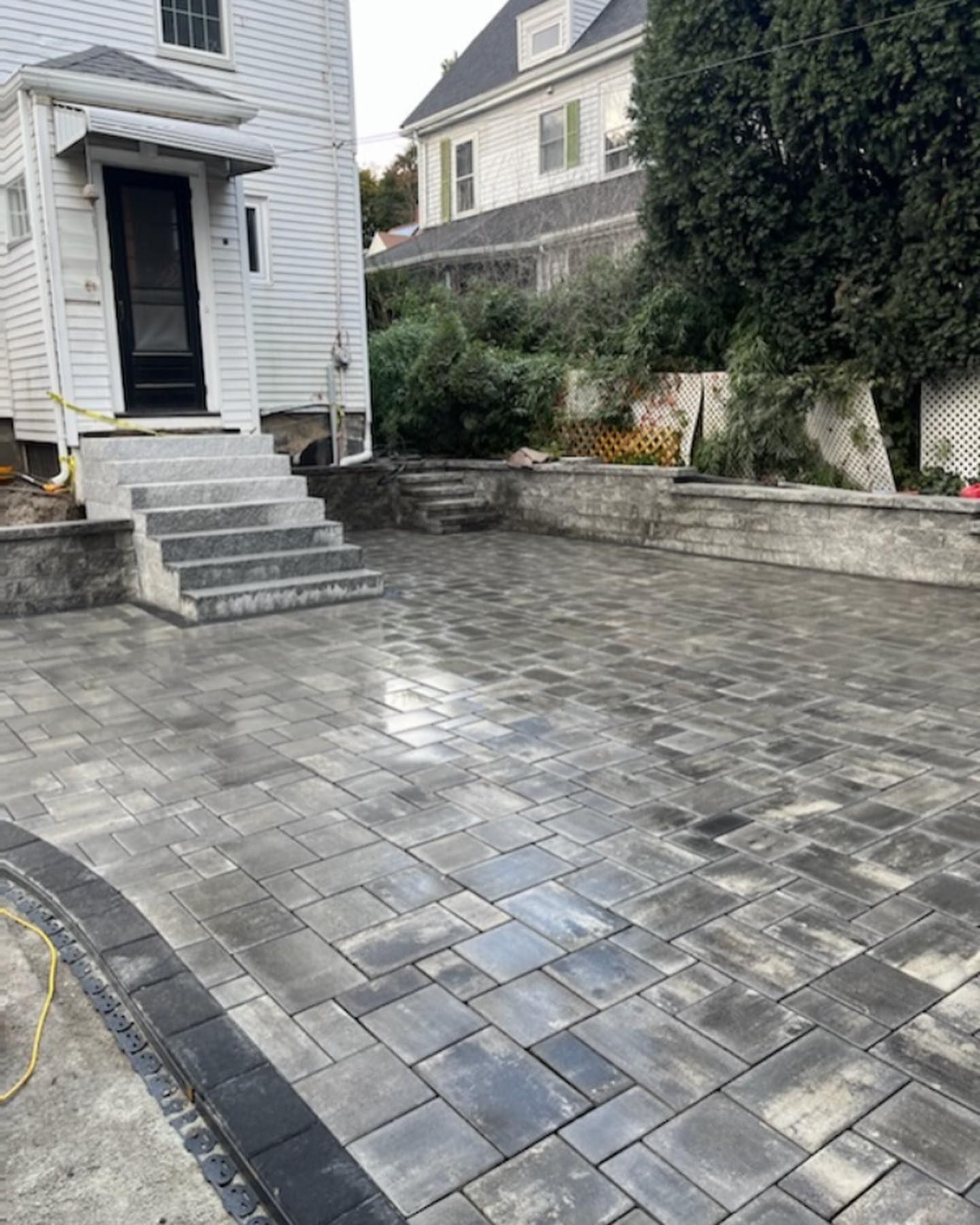 Paver patio with gray and black brickwork, concrete stairs leading to a white building.