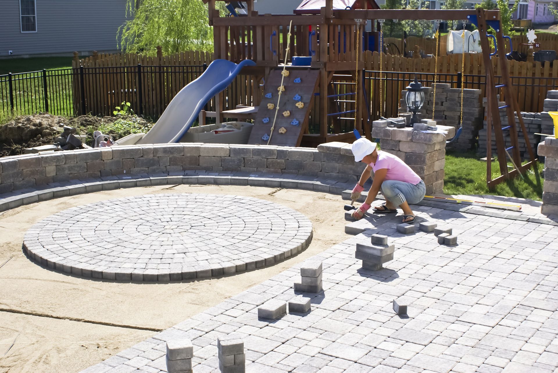 Person laying pavers in a backyard with a playground; circular patio being built.