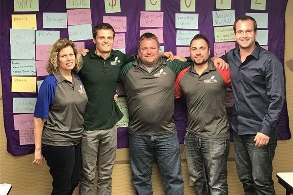 Five people standing arm-in-arm in front of a wall covered with colorful sticky notes