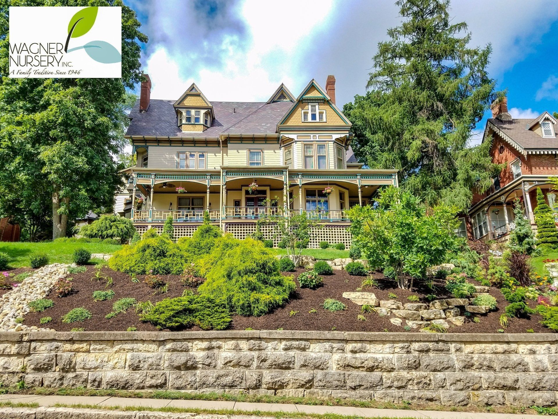 Large Victorian house with a wraparound porch, lush garden, and stone retaining wall under a blue sky