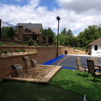 Backyard putting green with water features, patio chairs, and stone retaining wall under cloudy sky