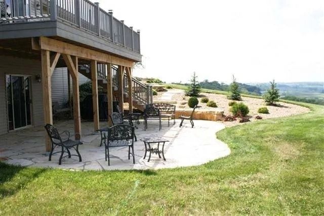 Patio with black metal tables and chairs beside a wooden deck, overlooking a grassy landscape and fields