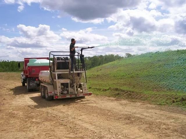 Worker on a farm machine spraying a green hillside beside a dirt road with a truck nearby