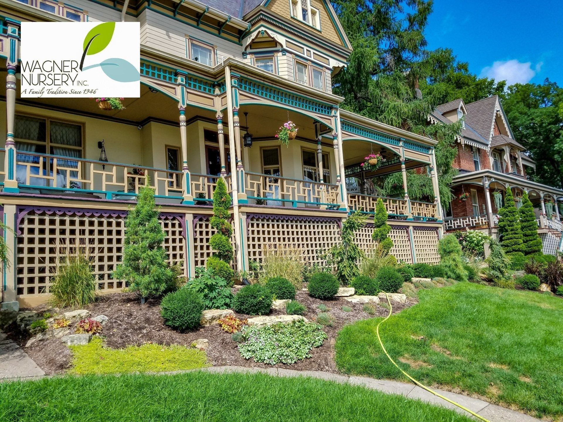 Victorian house with ornate porch, green trim, and landscaped garden under a bright blue sky