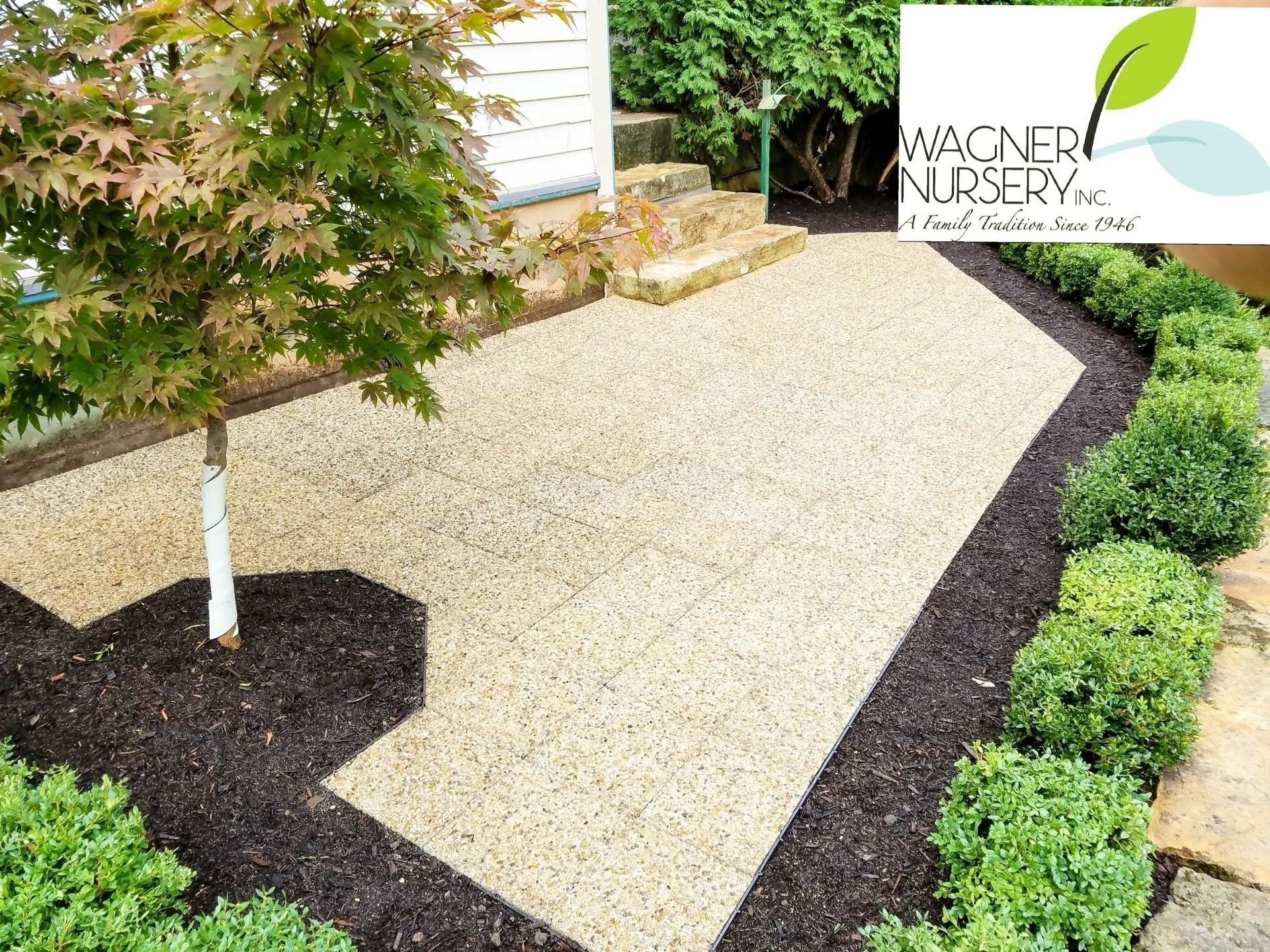 Gravel garden path bordered by dark mulch and shrubs beside a house and steps, with Wagner Nursery logo