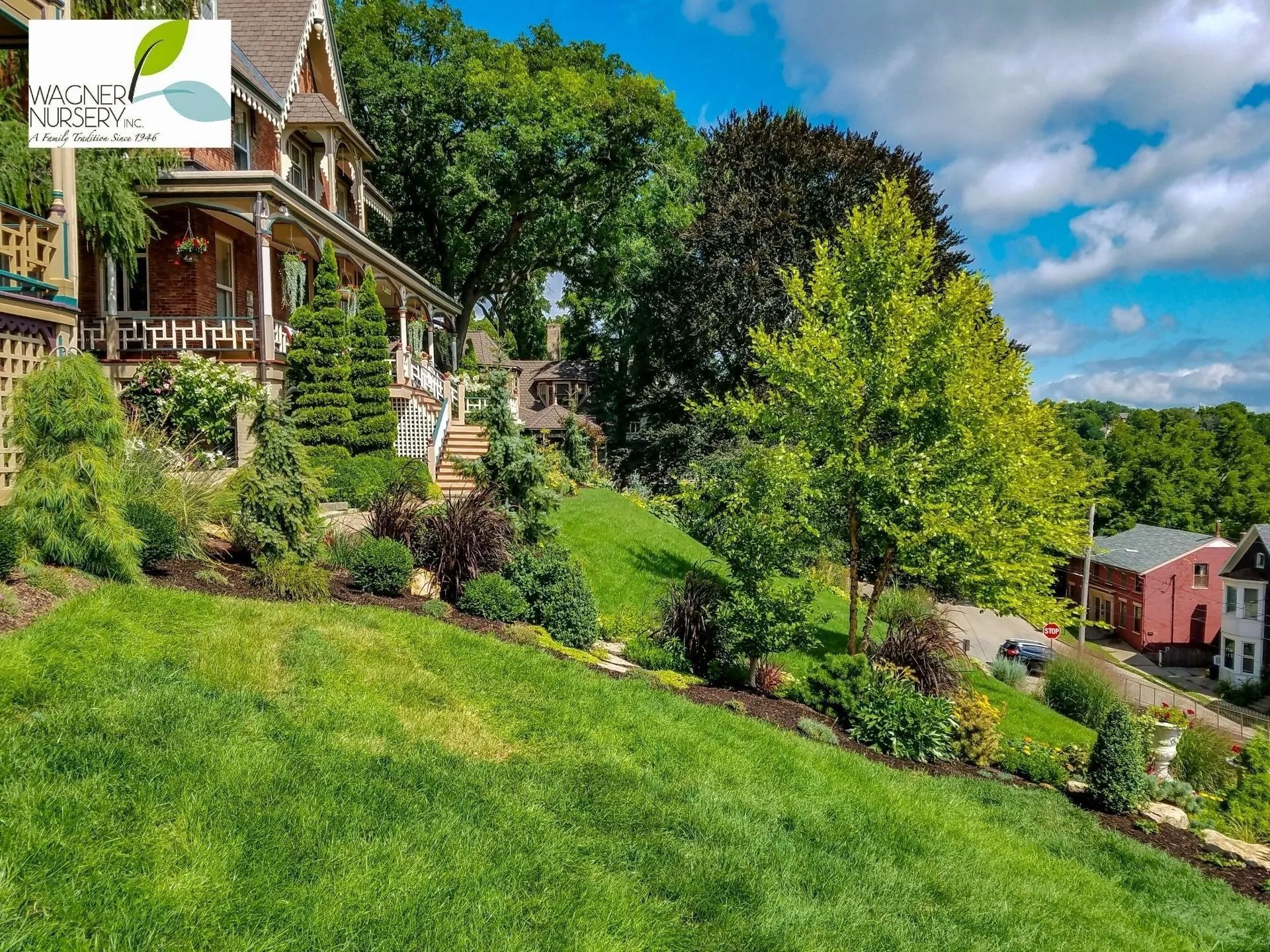 Sunlit hillside with a Victorian house, lush green lawn, trees, and a small red building under a blue sky.