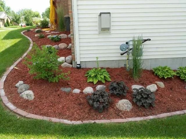 Curved front yard garden bed with red mulch, small shrubs, and decorative stones beside a white house