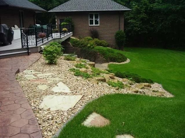 Backyard garden with stone path, gravel bed, trimmed shrubs, green lawn, and brick house patio
