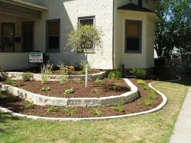 Curved flower bed with mulch and young tree beside a stucco house and lawn