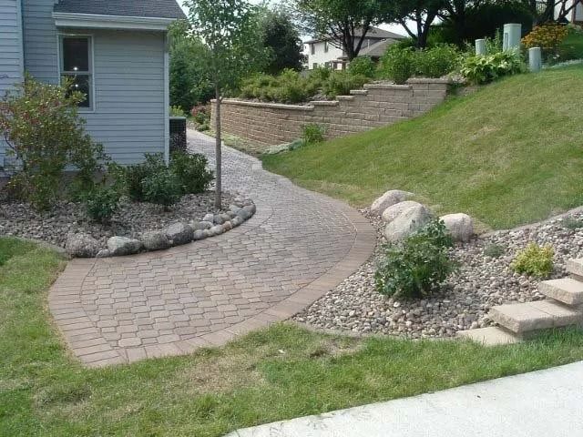 Curved brick walkway beside a gray house and landscaped grassy slope with rocks and shrubs