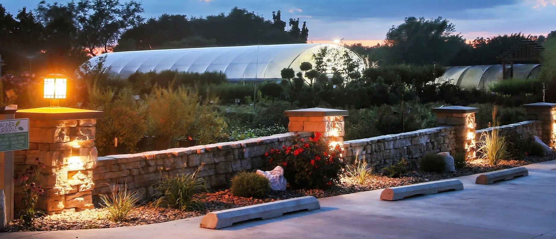 Lit stone entrance with lanterns, flowers, and a white greenhouse at dusk