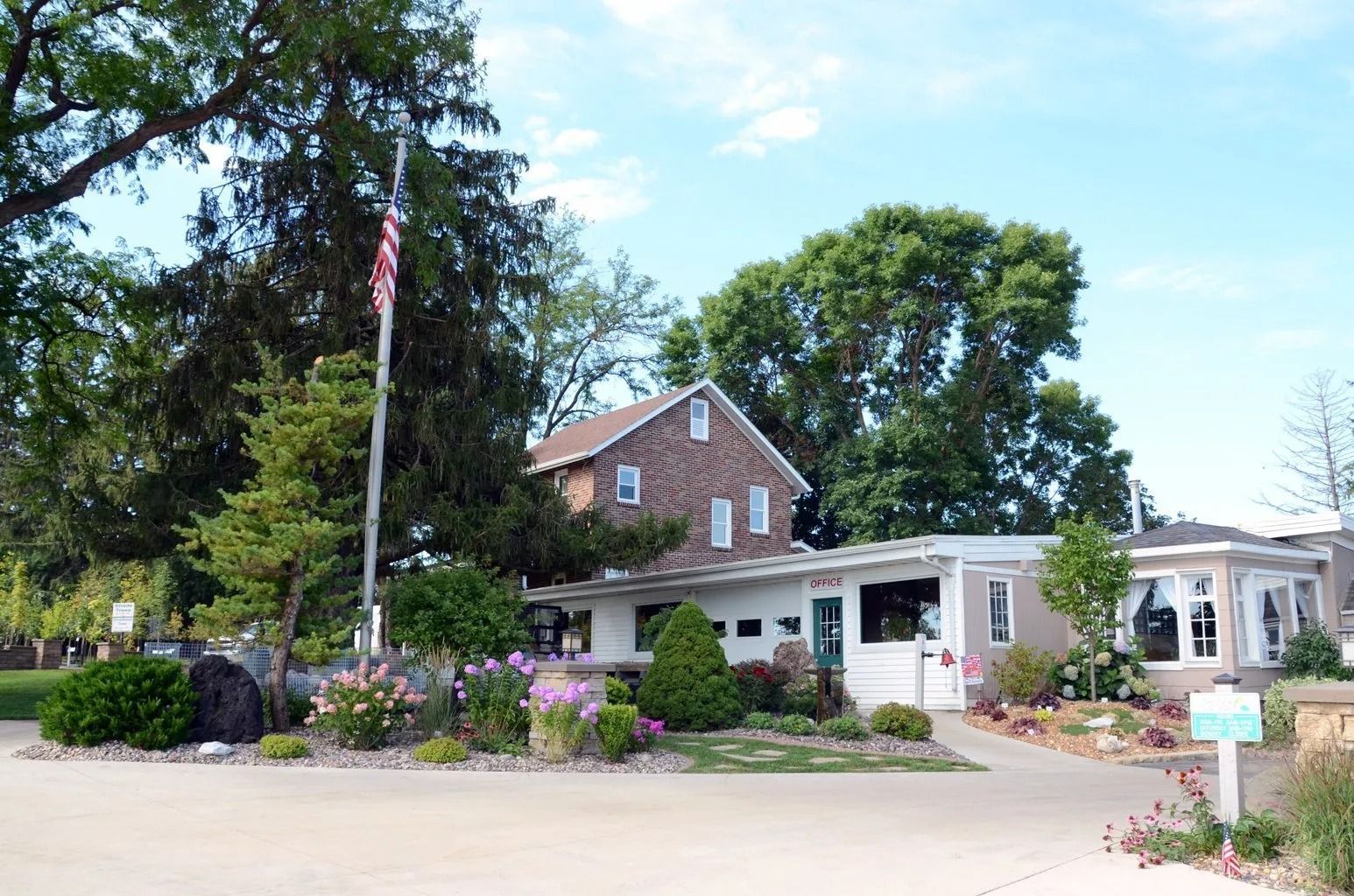 Brick house with white porch, garden flowers, and an American flag beside a driveway under trees