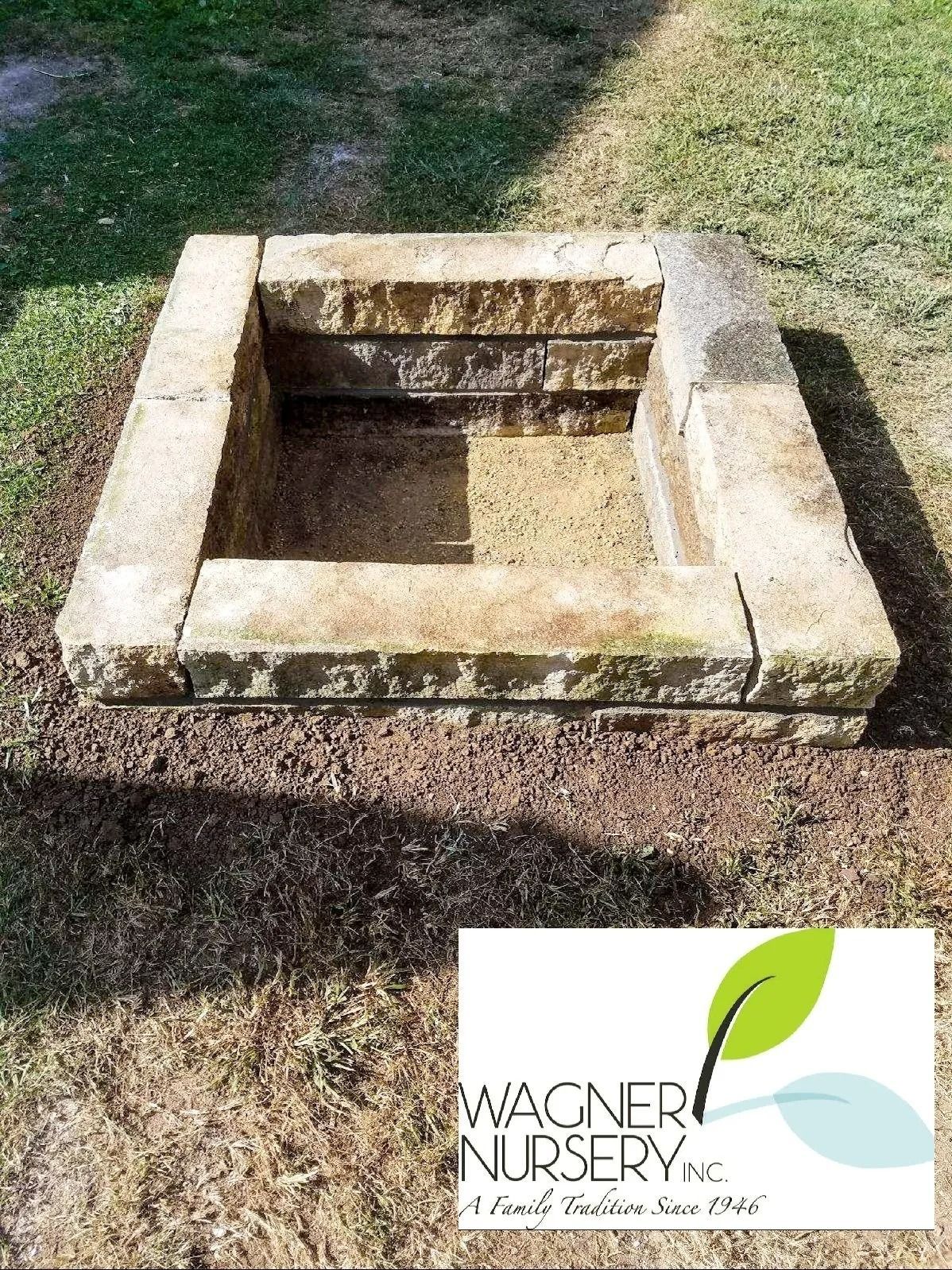 Stone outdoor fire pit with a square concrete border on grass, Wagners Nursery logo in the corner