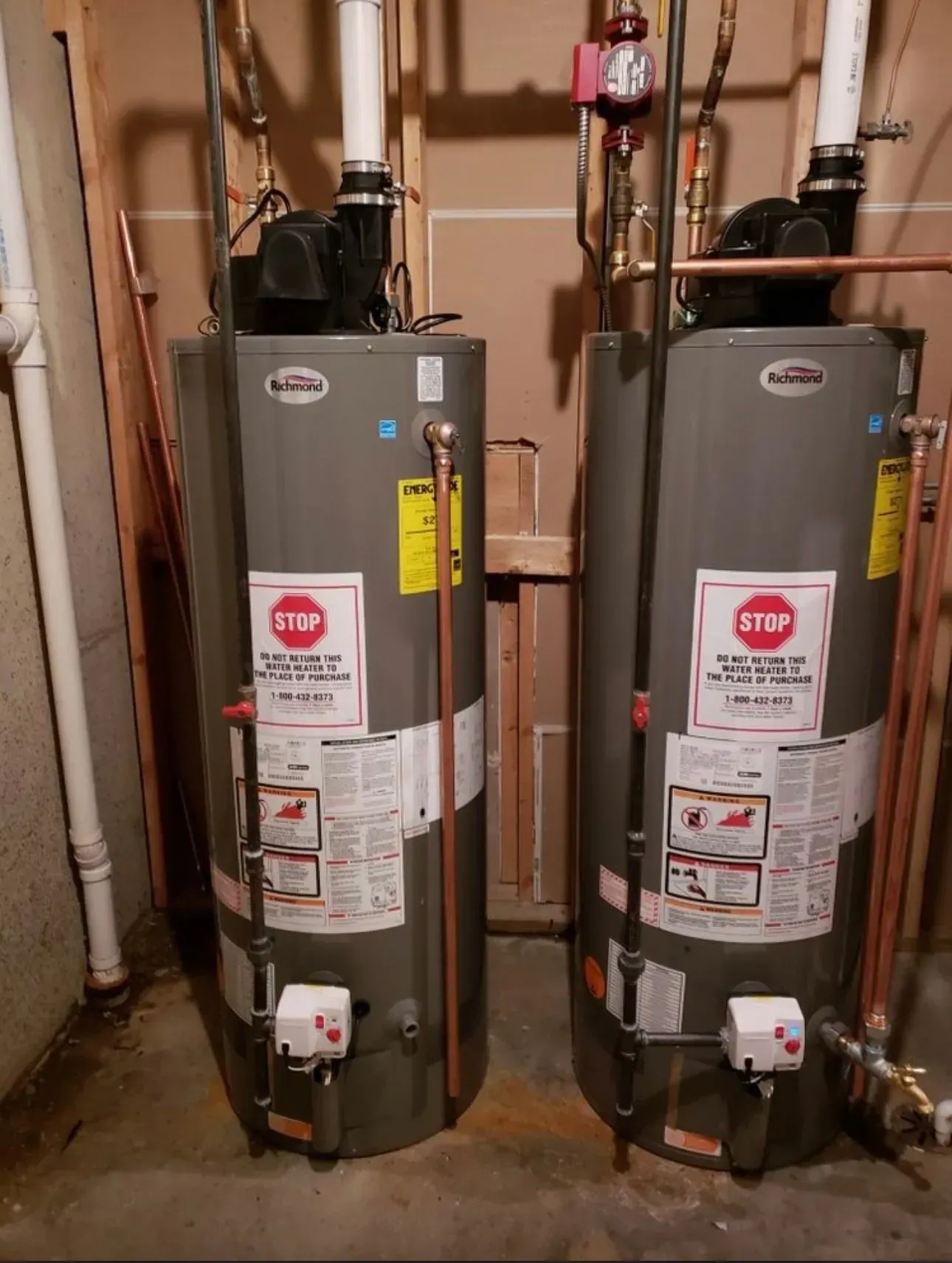 Two gray water heaters with copper pipes and white exhaust vents stand side by side in a basement.