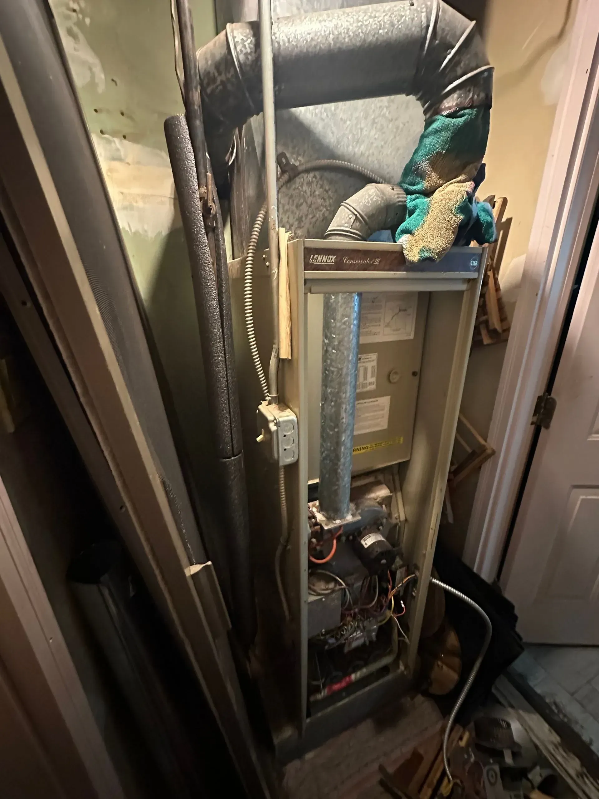 Furnace in a narrow utility room. Metal and components visible, ductwork above. Grey, beige, and brown colors.