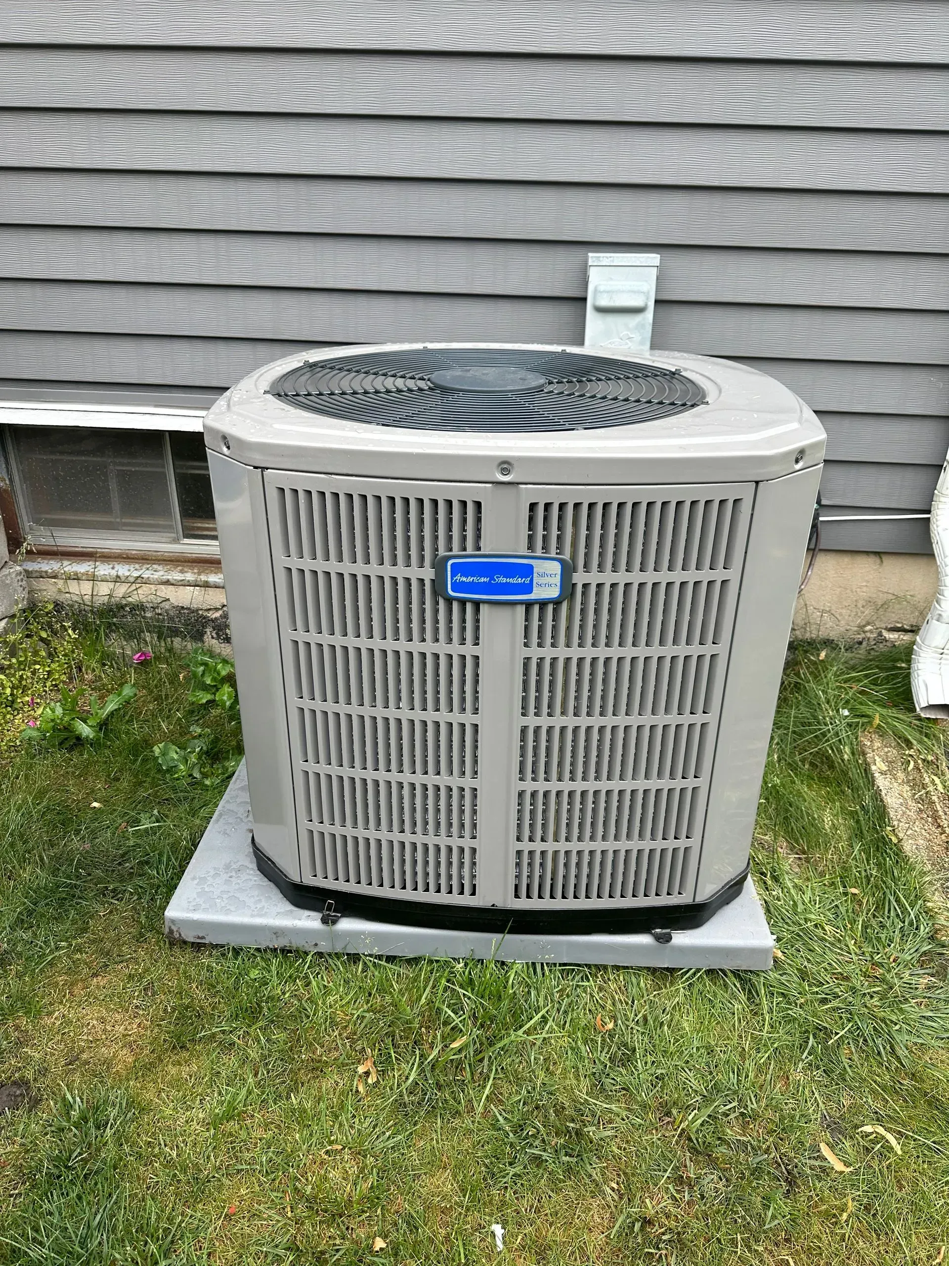 Air conditioner unit on a concrete pad, beige and gray, outdoors by a gray house wall.