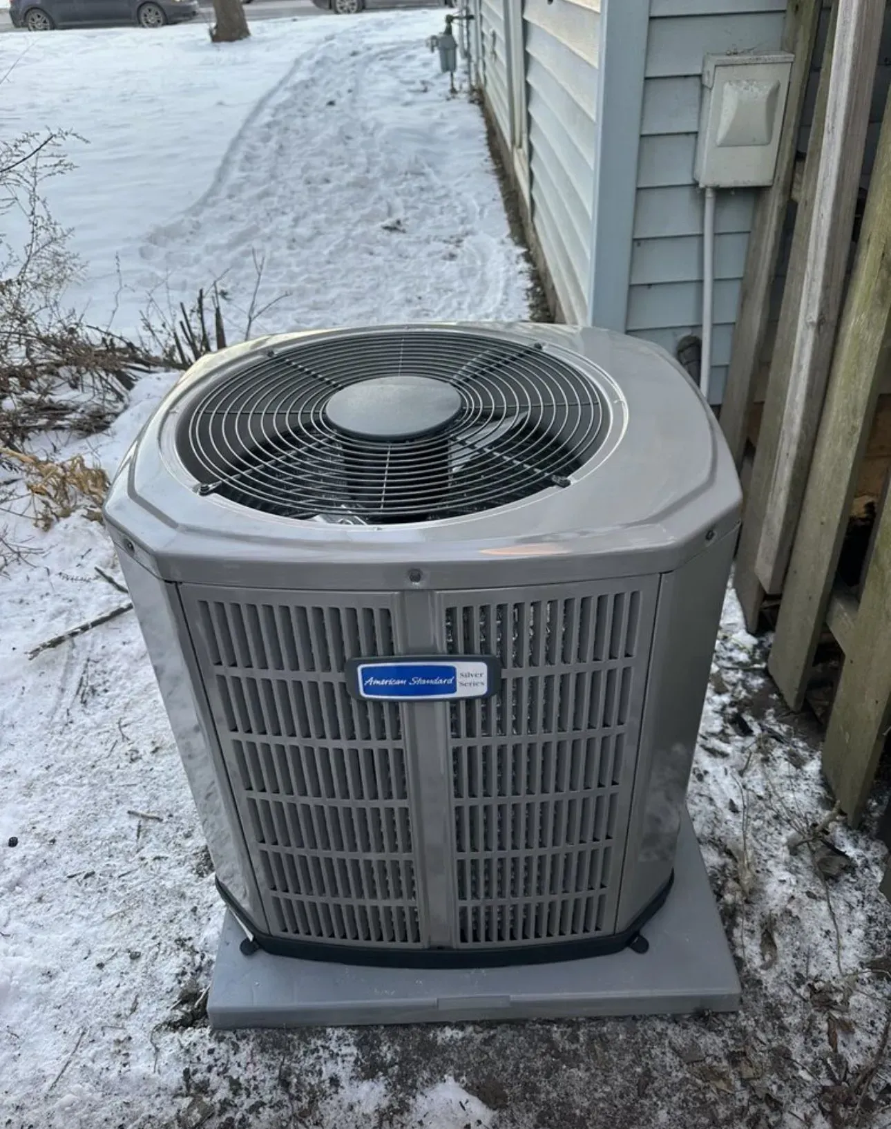 Gray air conditioning unit sits on a concrete pad in a snowy, outdoor setting.