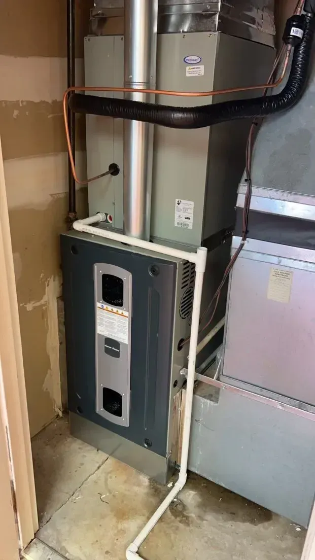 Furnace system in a utility room; gray and silver unit with white pipes and black ducting.