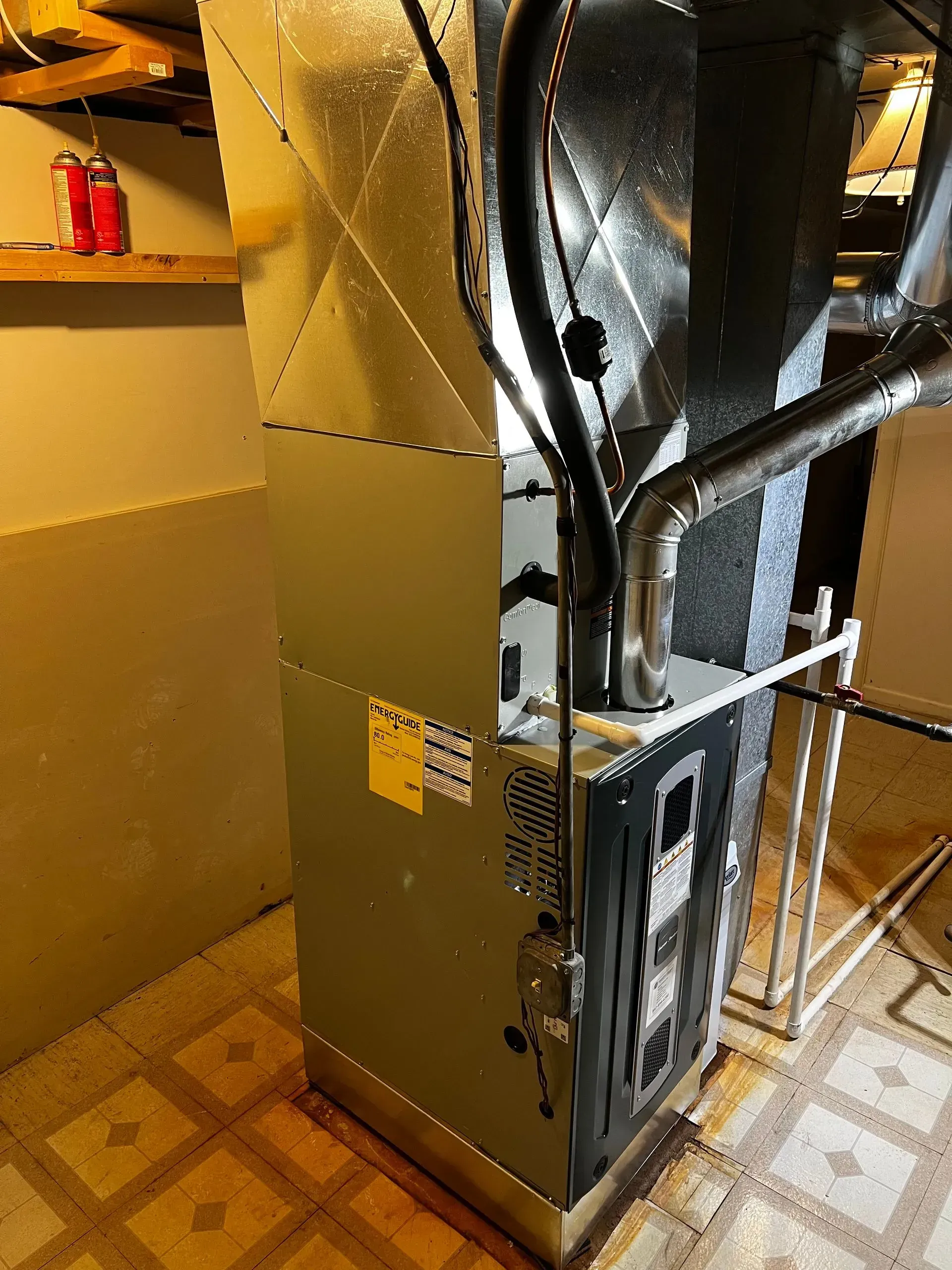 Furnace in a basement setting. Metal box with vents and pipes. Yellow tag on the side.