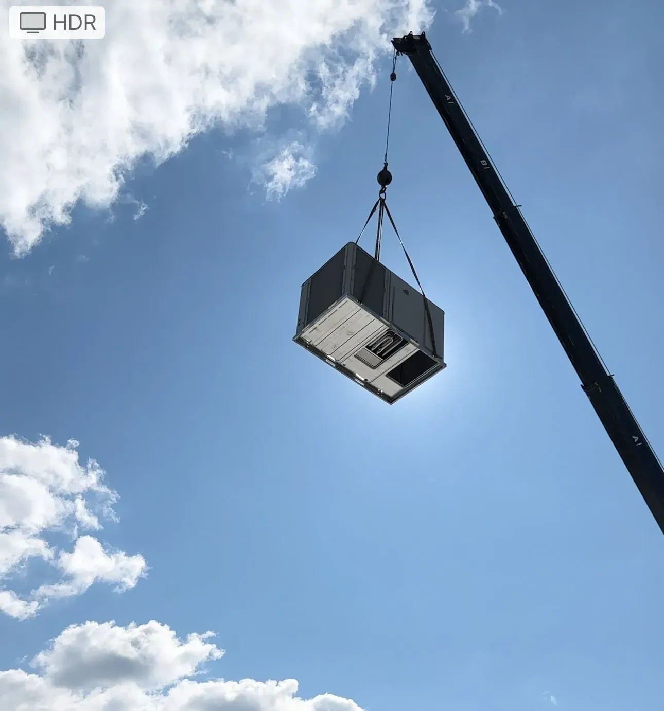 A large air conditioning unit suspended by a crane against a bright blue sky.