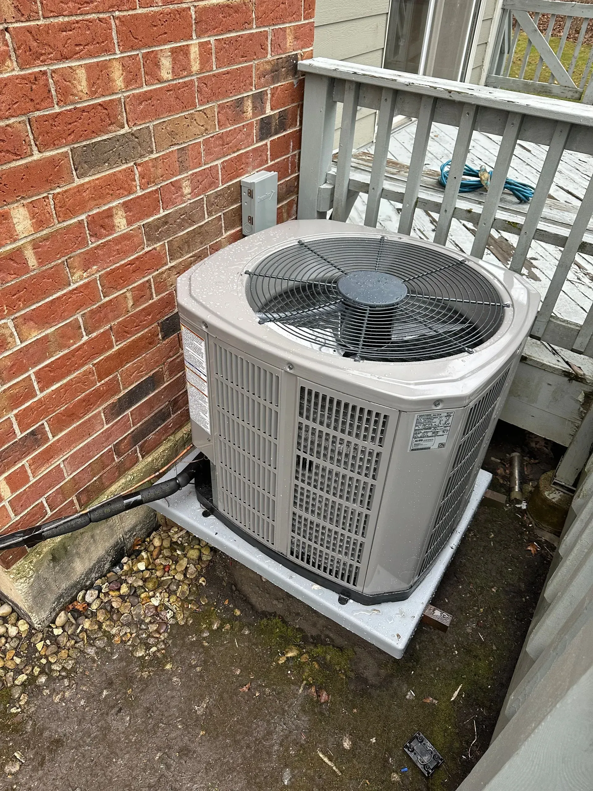 Outdoor air conditioning unit next to a brick wall and wooden deck.