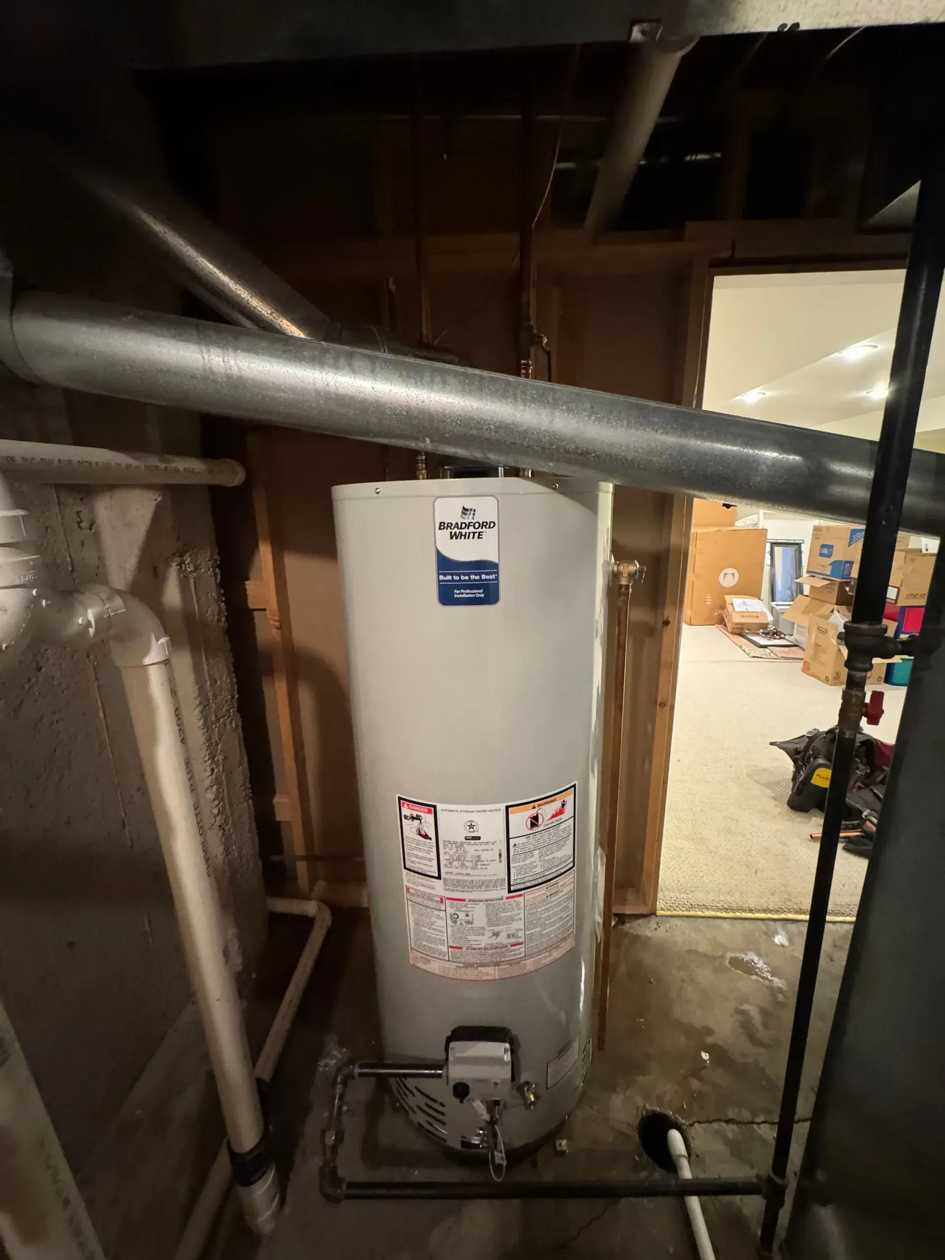 A natural gas water heater in a basement setting, with metal pipes and ductwork visible.