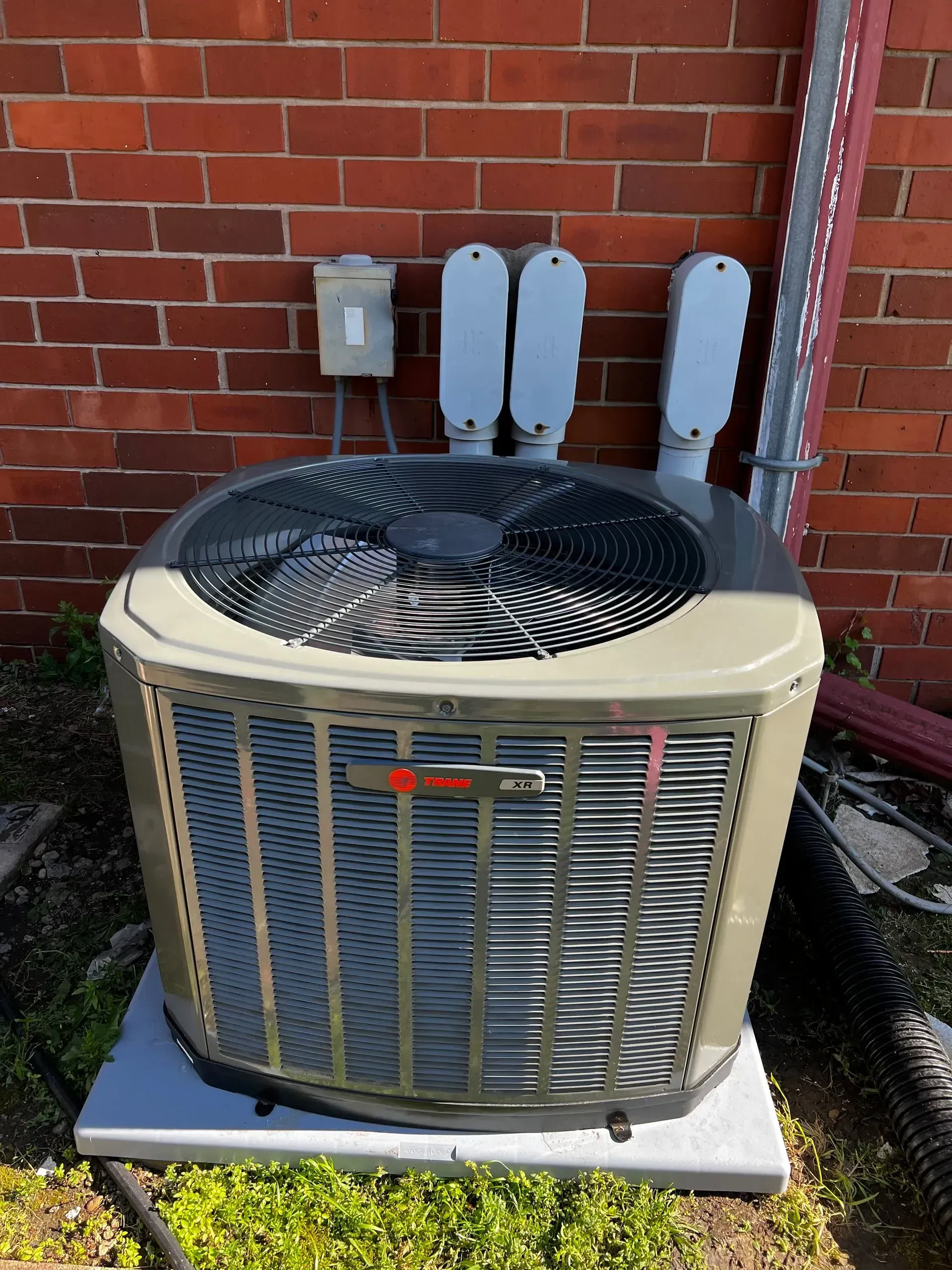 Outdoor Trane air conditioning unit against a red brick wall, with electrical conduits above.