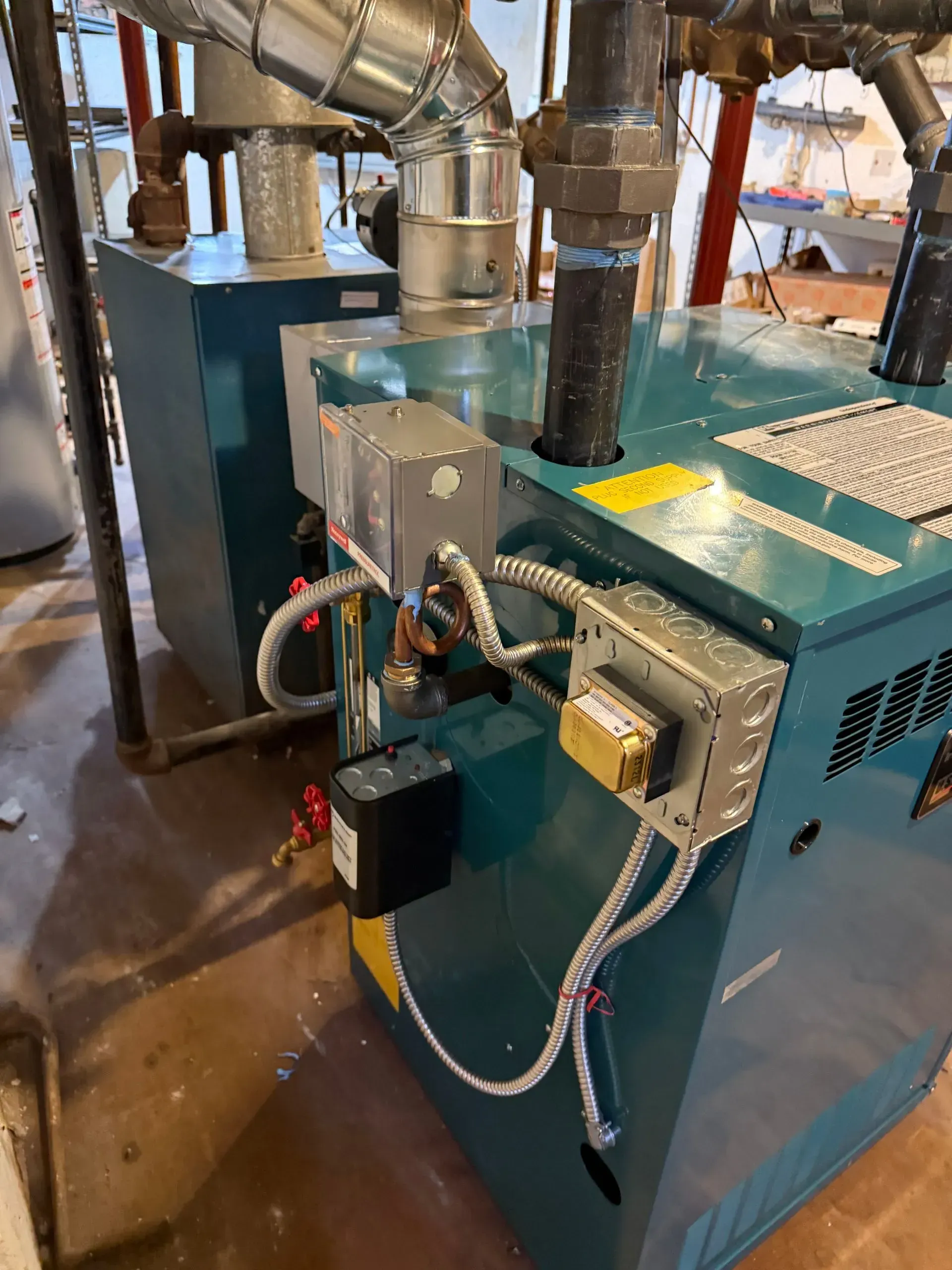 Two teal heating boilers in a basement setting, with silver pipes and electrical components visible.