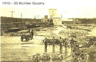 Men working at the Bichler Quarry, circa 1910-1920. Workers stand near carts and stone piles. Building visible in the background.