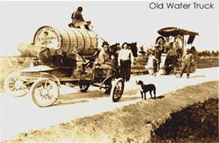 Old water truck with large barrel, several people, dog, and a horse-drawn cart on a dirt road.