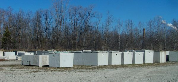 A row of concrete rectangular boxes sits in a gravel lot, trees in the background, blue sky.