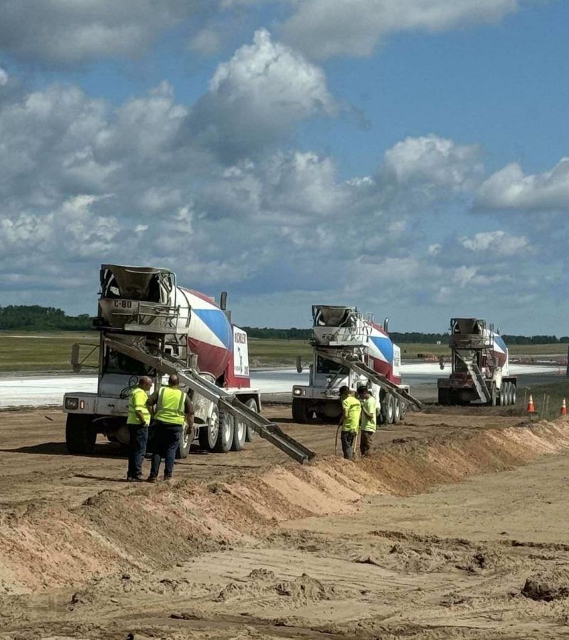 Three cement trucks pouring concrete on a construction site; workers in safety vests; blue sky with clouds.