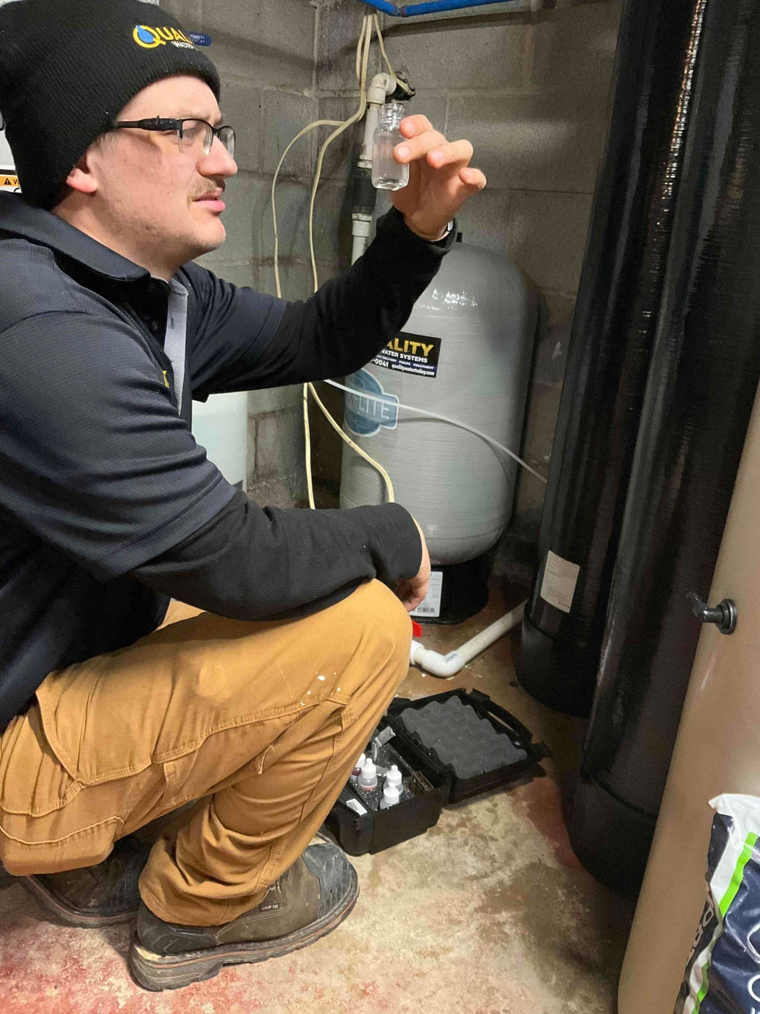 A technician wearing a beanie and work clothes examines a water sample in a vial in a basement with water treatment tanks.