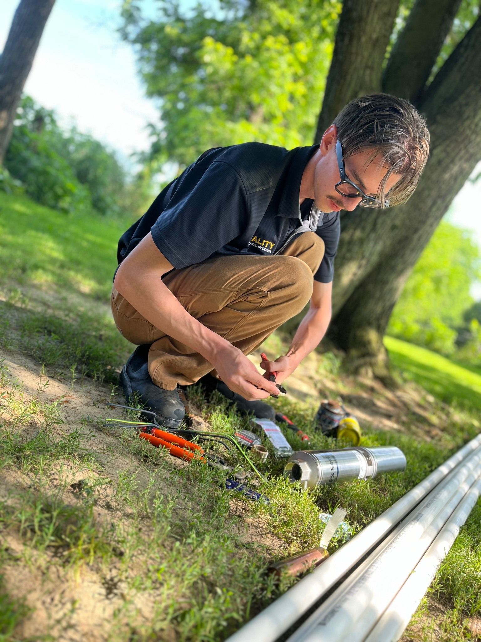 A person crouches outdoors, working on metal pipes and wires on a grassy patch near a tree.