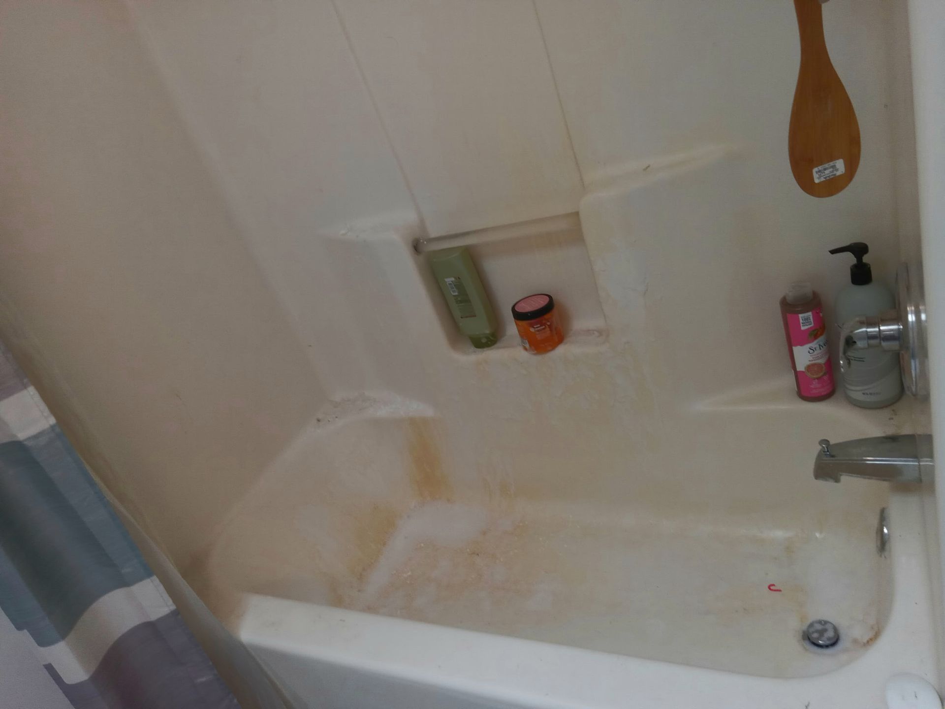 A white bathtub with orange stains, holding toiletries and a wooden brush, beside a blue and white shower curtain.